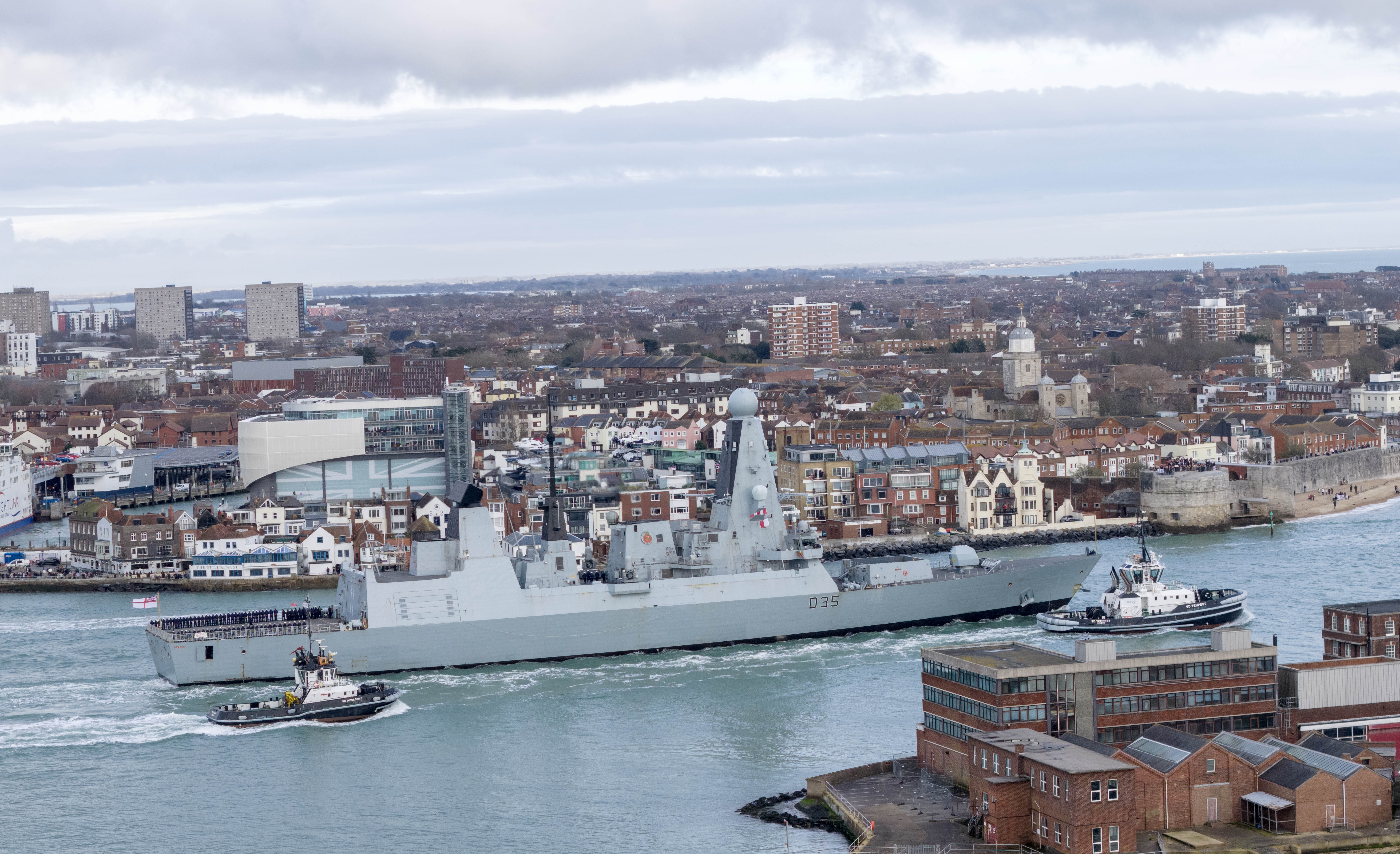 HMS Dragon, a Type 45 destroyer with the pennant number D35, departing Portsmouth Harbour assisted by two tugboats.