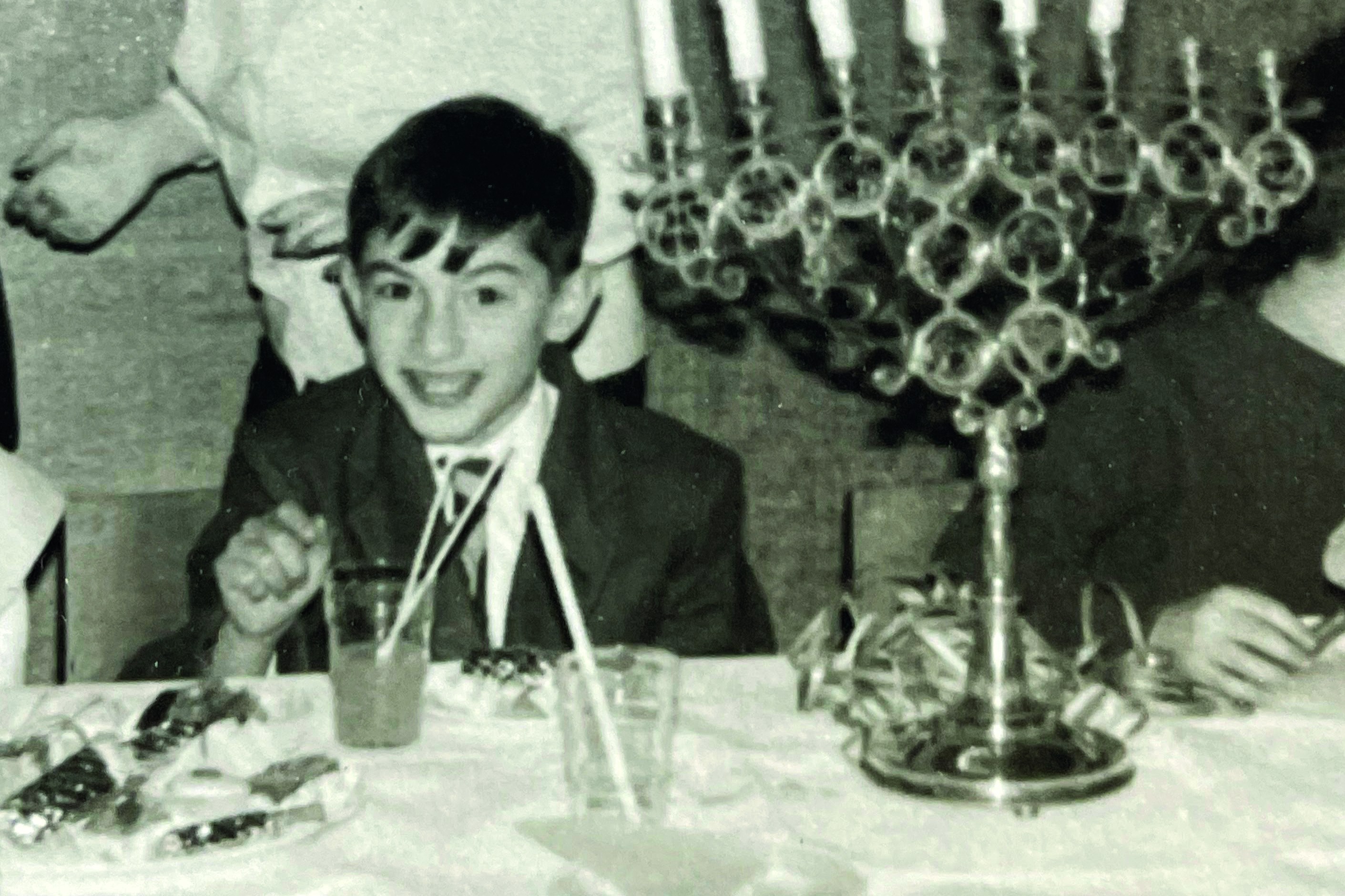 A grayscale photo shows a boy at a Chanukah party, seated at a table with a menorah with candles, and a woman standing behind him.