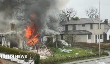 House in flames on a street - billowing grey smoke rises from the single-storey white building