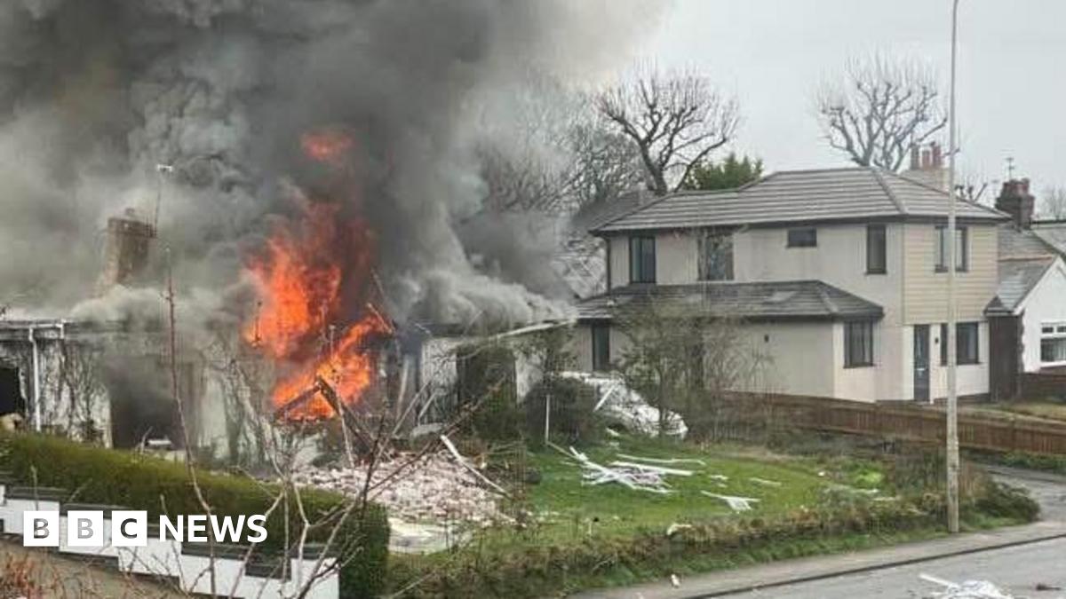 House in flames on a street - billowing grey smoke rises from the single-storey white building