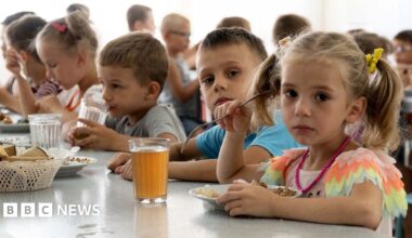 Children from an orphanage in the Donetsk region, eat a meal at a camp in Zolotaya Kosa, the settlement on the Sea of Azov, Rostov region, southwestern Russia, Friday, July 8, 2022. A little girl with plaits is on the right, closest to the camera, she is looking sadly at the camera and holding a fork in her right hand over her plate of food. A glass of red-brown liquid is on the table beside her. A boy next to her is also looking at the camera. Other children are busy eating and drinking.
