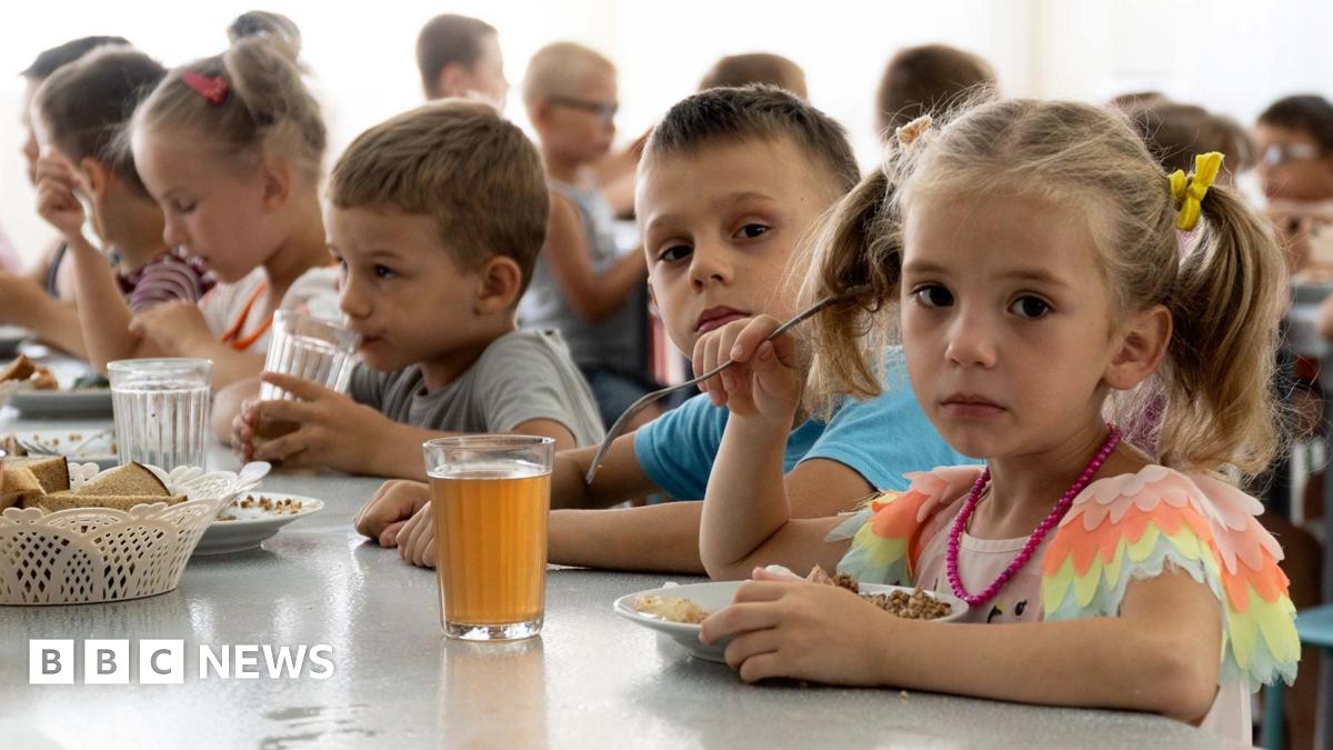 Children from an orphanage in the Donetsk region, eat a meal at a camp in Zolotaya Kosa, the settlement on the Sea of Azov, Rostov region, southwestern Russia, Friday, July 8, 2022. A little girl with plaits is on the right, closest to the camera, she is looking sadly at the camera and holding a fork in her right hand over her plate of food. A glass of red-brown liquid is on the table beside her. A boy next to her is also looking at the camera. Other children are busy eating and drinking.