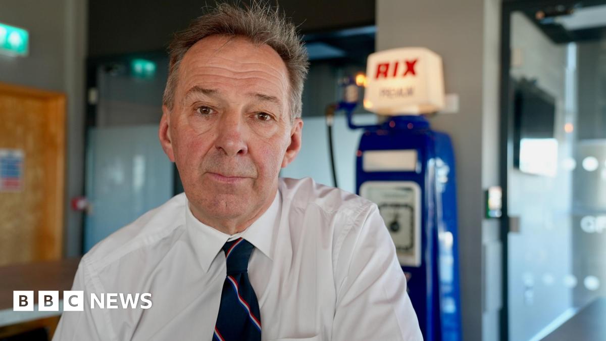 Duncan Lambert, wearing a white shirt and a blue tie sits in a room in front of an old Rix Petroleum petrol pump