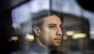 A man with short dark hair looks contemplatively through a reflective surface.