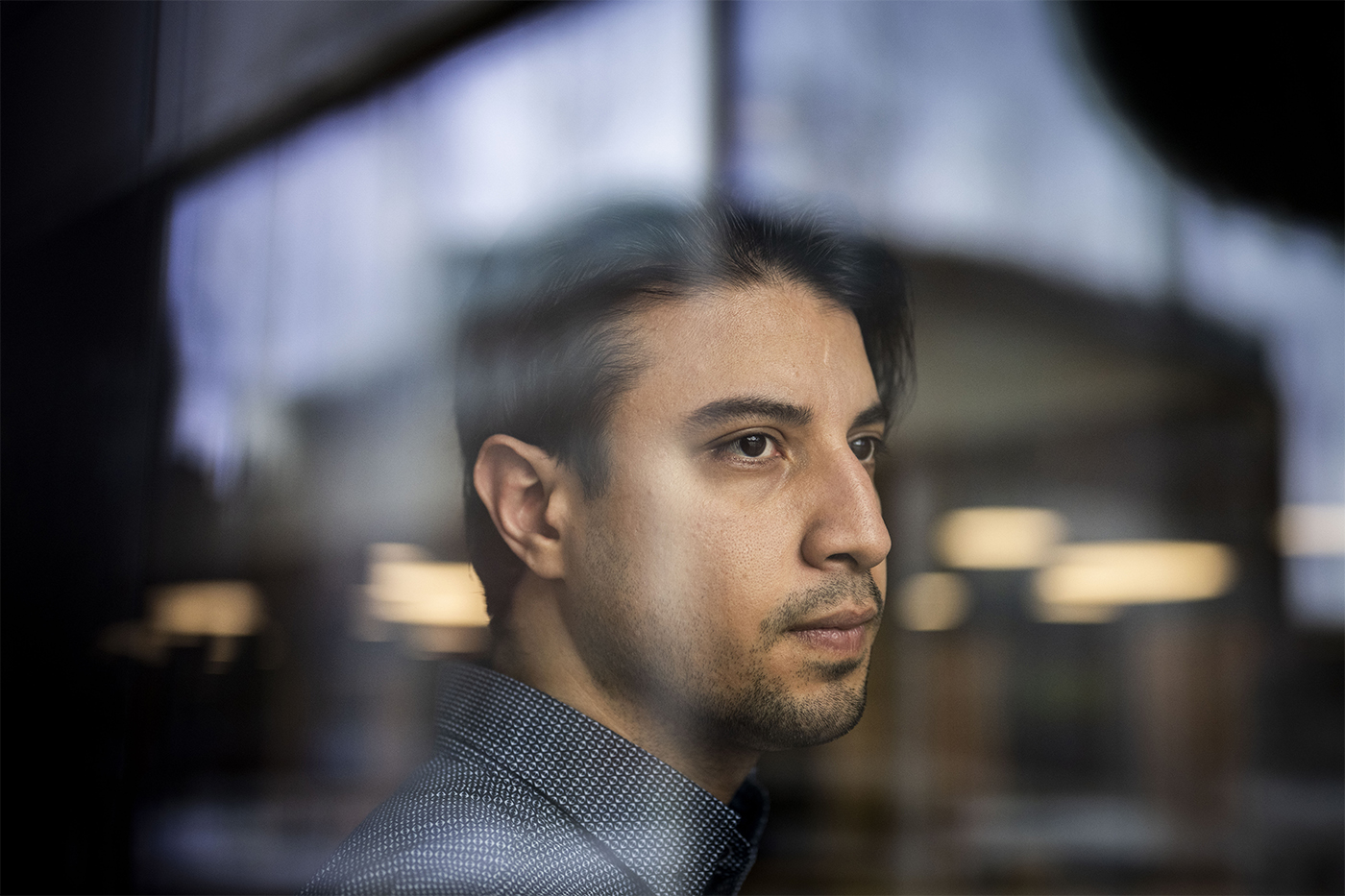 A man with short dark hair looks contemplatively through a reflective surface.