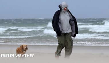 Man and his dog walking along a beach where sand is visibily lifted up with large waves in the background