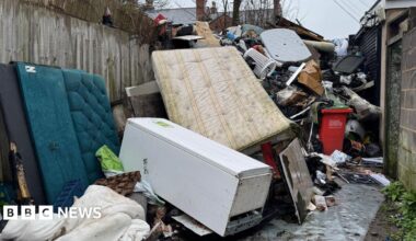 An alleyway between a concrete wall and garden fences. The alleyway is full of dumped rubbish including mattresses, fridges, sofas, duvets and other household waste