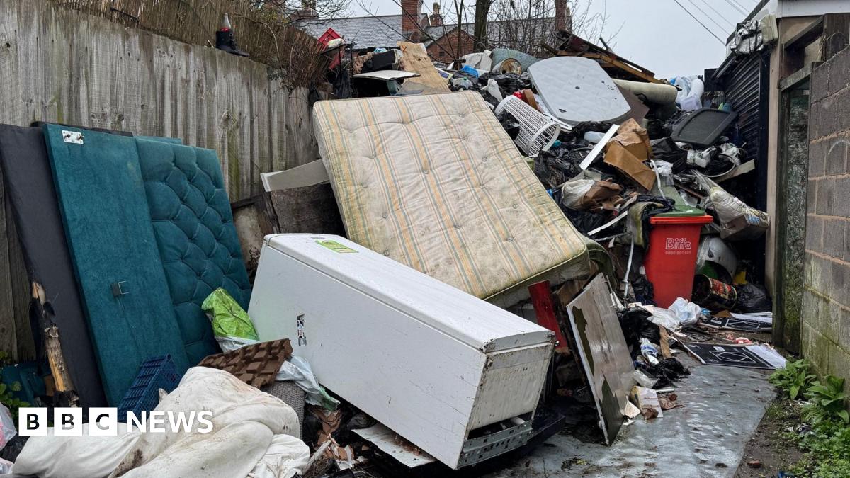 An alleyway between a concrete wall and garden fences. The alleyway is full of dumped rubbish including mattresses, fridges, sofas, duvets and other household waste