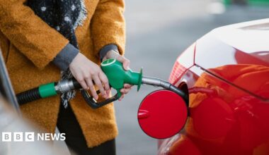 Close up of a woman in an ochre coloured warm coat inserting a petrol nozzle into a bright red car petrol tank.