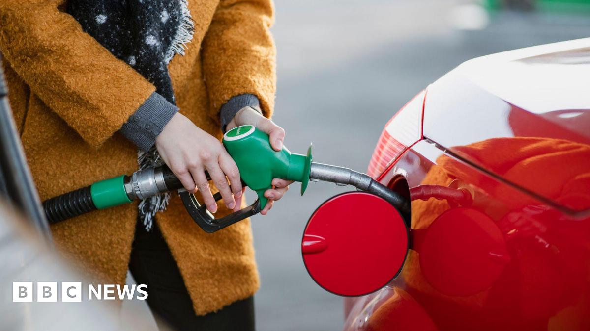 Close up of a woman in an ochre coloured warm coat inserting a petrol nozzle into a bright red car petrol tank.