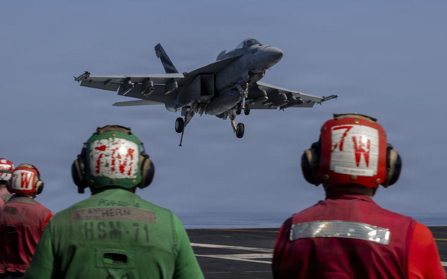 A military fighter jet lands on an aircraft carrier, as members of the flight crew, seen from behind, observe in the foreground.