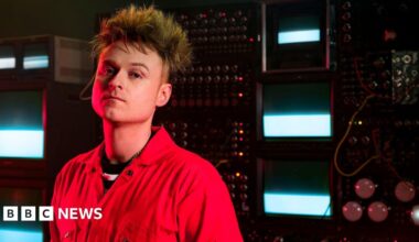 Look Mum No Computer, aka musician Sam Battle, stands in front of a bank of vintage synthesisers and TV monitors.