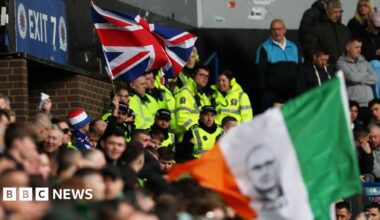 A woman with dark hair and a multiple colour jumper siting on a green sofa inside her living room.