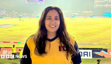 Yasmin stands at a sports game, in a yellow sports top, with a wide smile. The pitch can be seen in the background