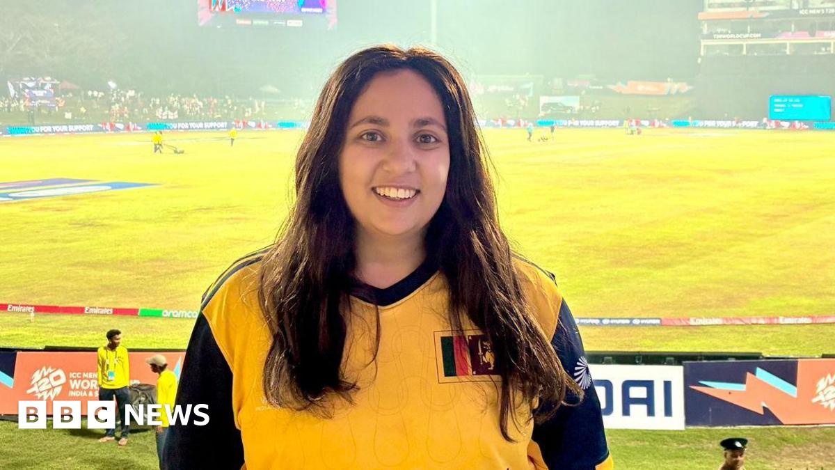 Yasmin stands at a sports game, in a yellow sports top, with a wide smile. The pitch can be seen in the background