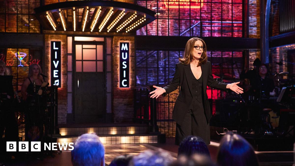 Tina Fey on the set of Saturday Night Live UK with a sign saying live music behind her - a band can be seen on the stage, and audience members are in front of her