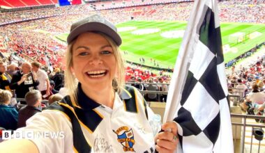 A woman holding a black and white racing flag and a football shirt stands in the stands of a large stadium. A football pitch can be seen behind her.