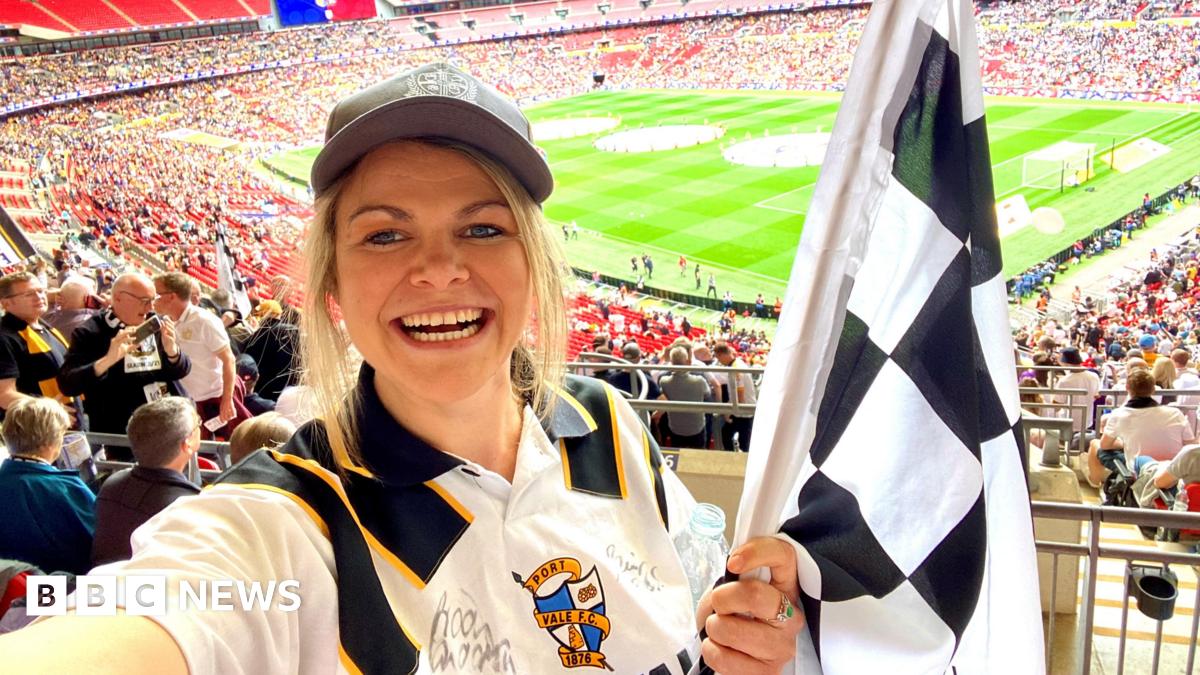 A woman holding a black and white racing flag and a football shirt stands in the stands of a large stadium. A football pitch can be seen behind her.
