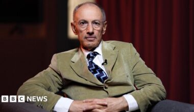 A man looking at the camera. He has a bald head but greying hair on the sides. He is sat in a grey armchair and has a green cord jacket on, with a white shirt underneath, and a dark blue and light blue zig-zag tie. The jacket is done up. He also has thin-rimmed glasses on. There is a burnt orange curtain in the background.