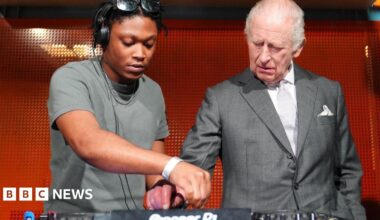Christian St Louis helps King Charles operate his DJ deck during a visit to Aviva Studios in Manchester, the home of Factory International. Charles is wearing a light grey suit with a white shirt and a pale grey tie.