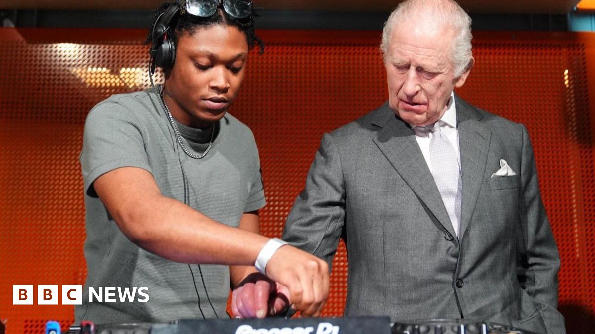 Christian St Louis helps King Charles operate his DJ deck during a visit to Aviva Studios in Manchester, the home of Factory International. Charles is wearing a light grey suit with a white shirt and a pale grey tie.