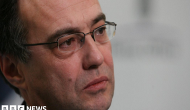 Phil Woolas, wearing a suit and glasses, looks thoughtful as his photo is taken by a professional photographer. He is photographed partly side on looking to the right of the frame.