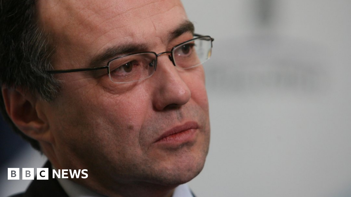 Phil Woolas, wearing a suit and glasses, looks thoughtful as his photo is taken by a professional photographer. He is photographed partly side on looking to the right of the frame.