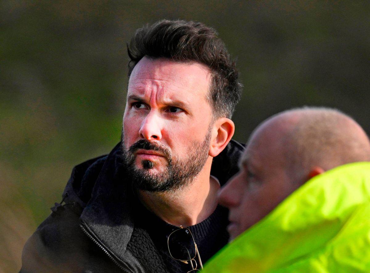 An individual with facial hair is engaged in conversation with another individual, both of them dressed in outdoor attire.