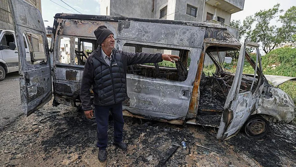 An elderly man stands next to a burnt-out van. 