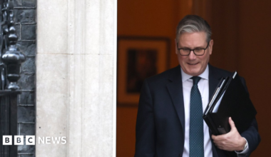 Prime Minister Keir Starmer departs Downing Street ahead of his statement on Iran. He wears a black suit, white shirt and navy tie and is carrying two black folders of documents