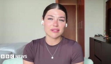 A woman with dark hair sits in a hotel room. She is wearing a purple t-shirt and silver chain around her neck.