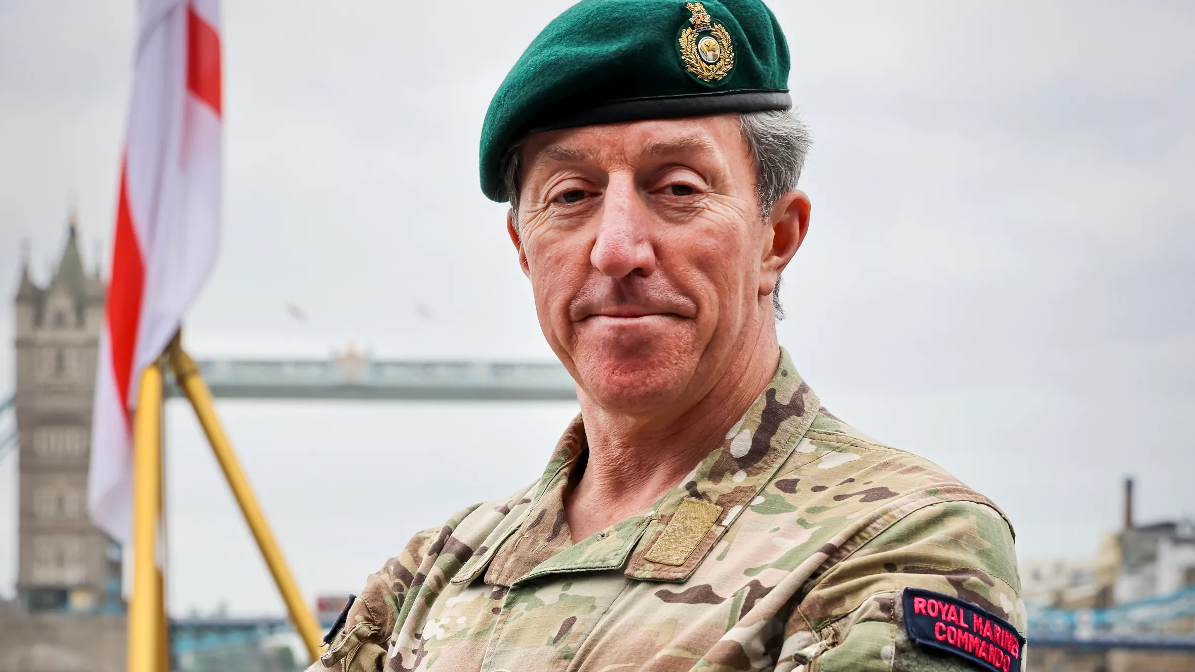 Portrait of General Sir Gwyn Jenkins, First Sea Lord, in military fatigues and green beret, standing with arms crossed on a ship deck with the Tower Bridge in the background.