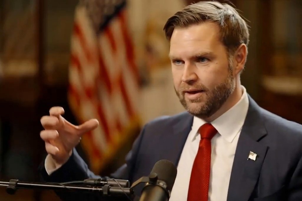 A man in a suit and red tie gestures with his hand during an interview, with an American flag out of focus in the background.
