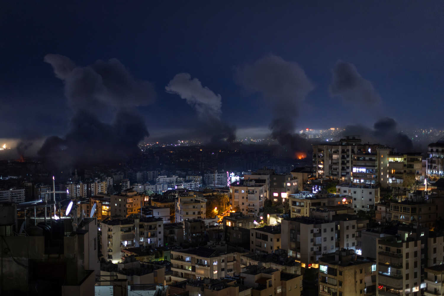 A city at night under a dark sky, many buildings lit. Thick, dark smoke columns rise from various points across the urban landscape.