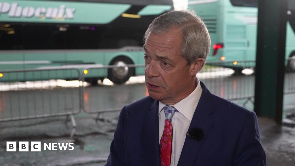 A man with grey hair, wearing a dark blue suit, white shirt and red tie. There are two green buses stationed behind him.