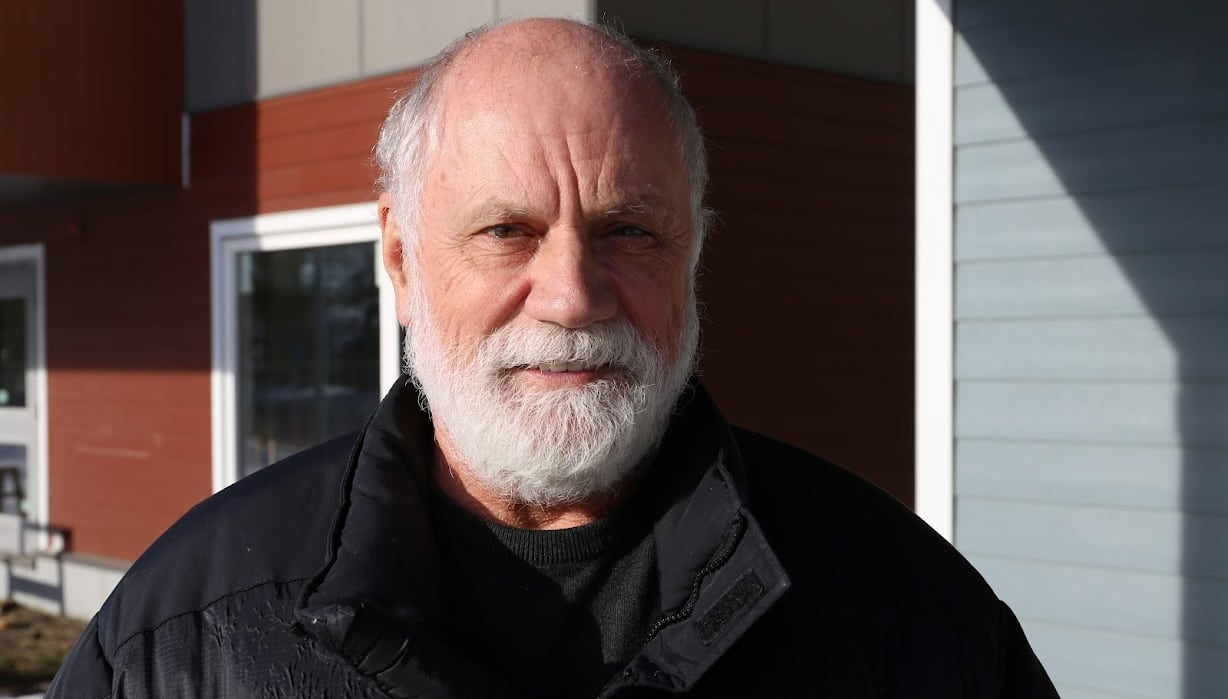 A balding man with white hair and a beard wears a black winter jacket.