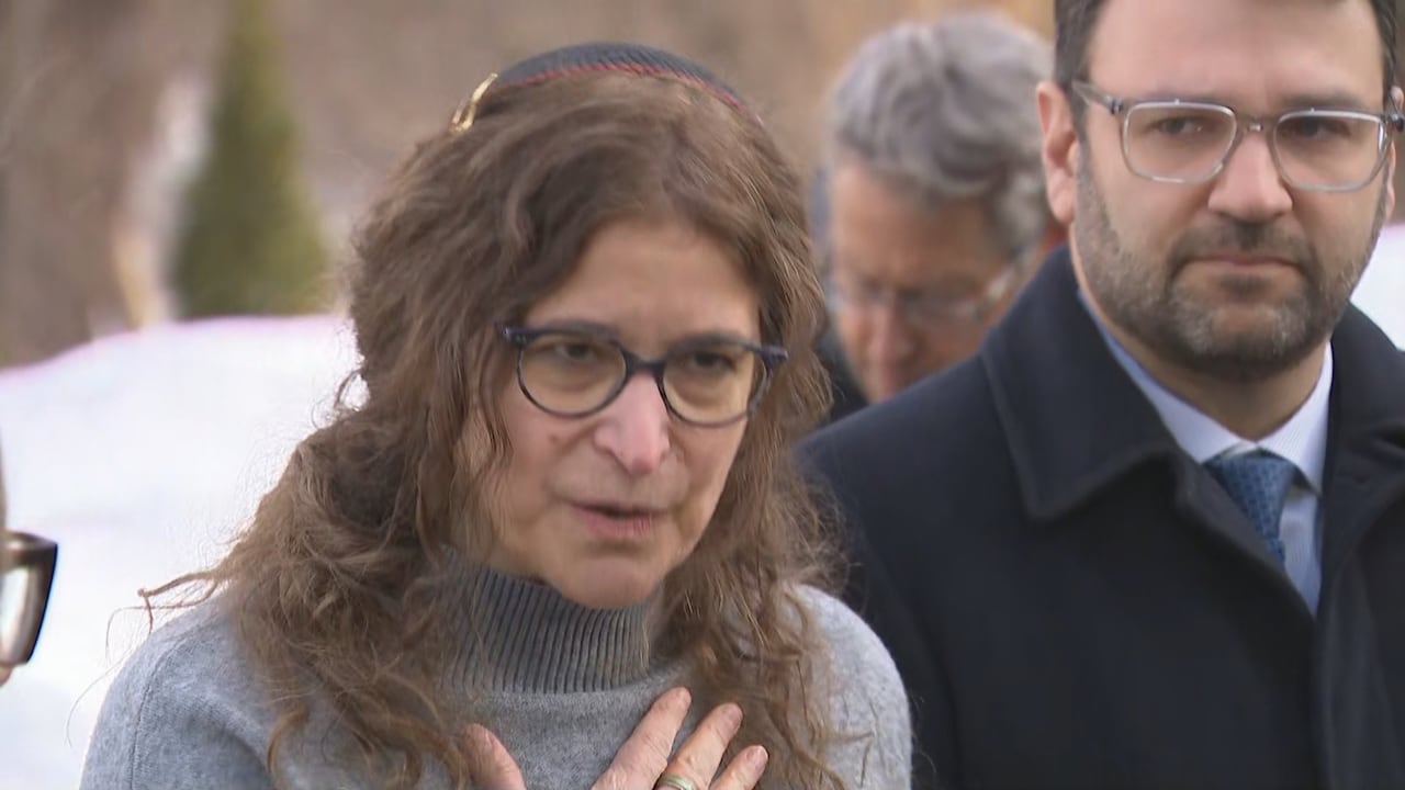 A middle-aged Rabbi holds her hand on her heart and speaks to someone off camera. SHe is standing outdoors in winter, next to a man in a suit and black overcoat