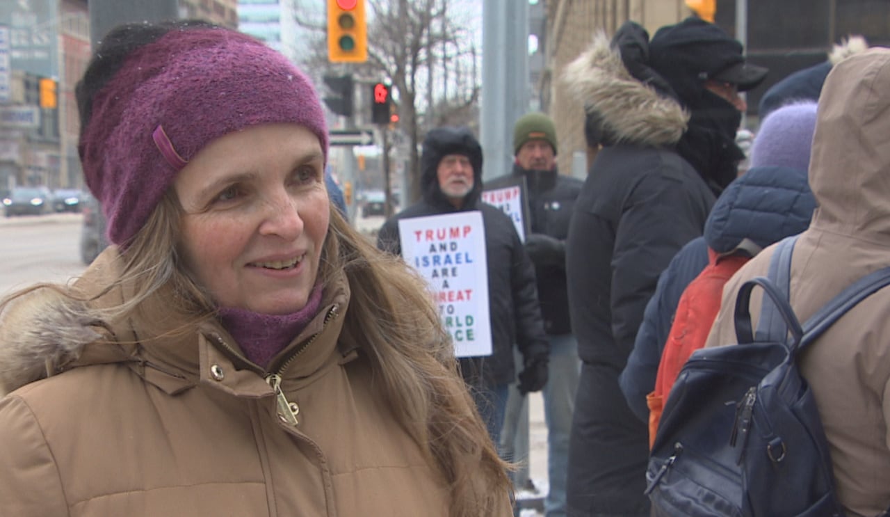 Woman wearing a brown winter jacket stands on a city street corner while people holding protest signs stand behind her