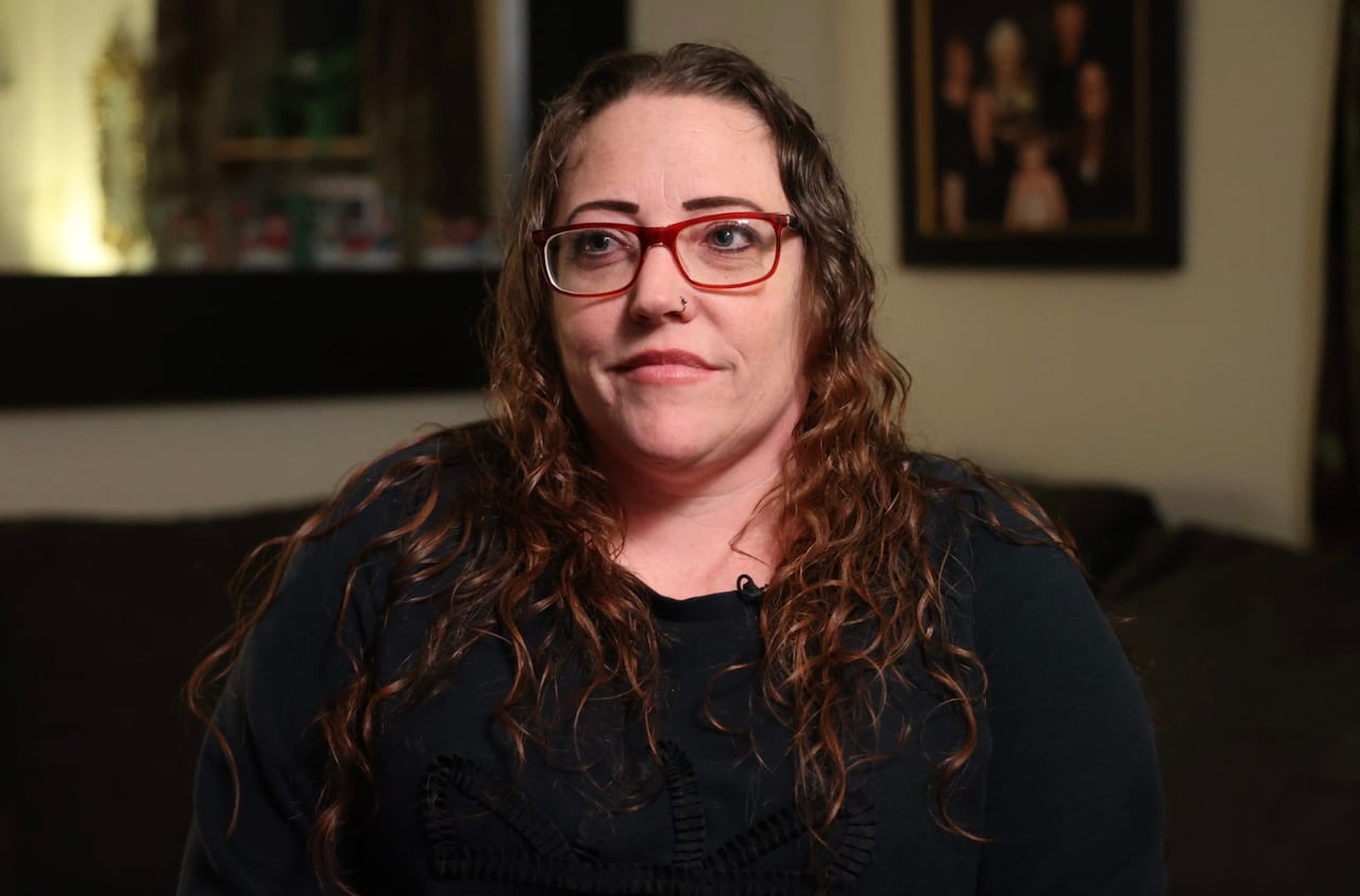 A woman with long, brown, curly hair sits in her living room. She's wearing a black long-sleeved shirt and red eyeglasses. 