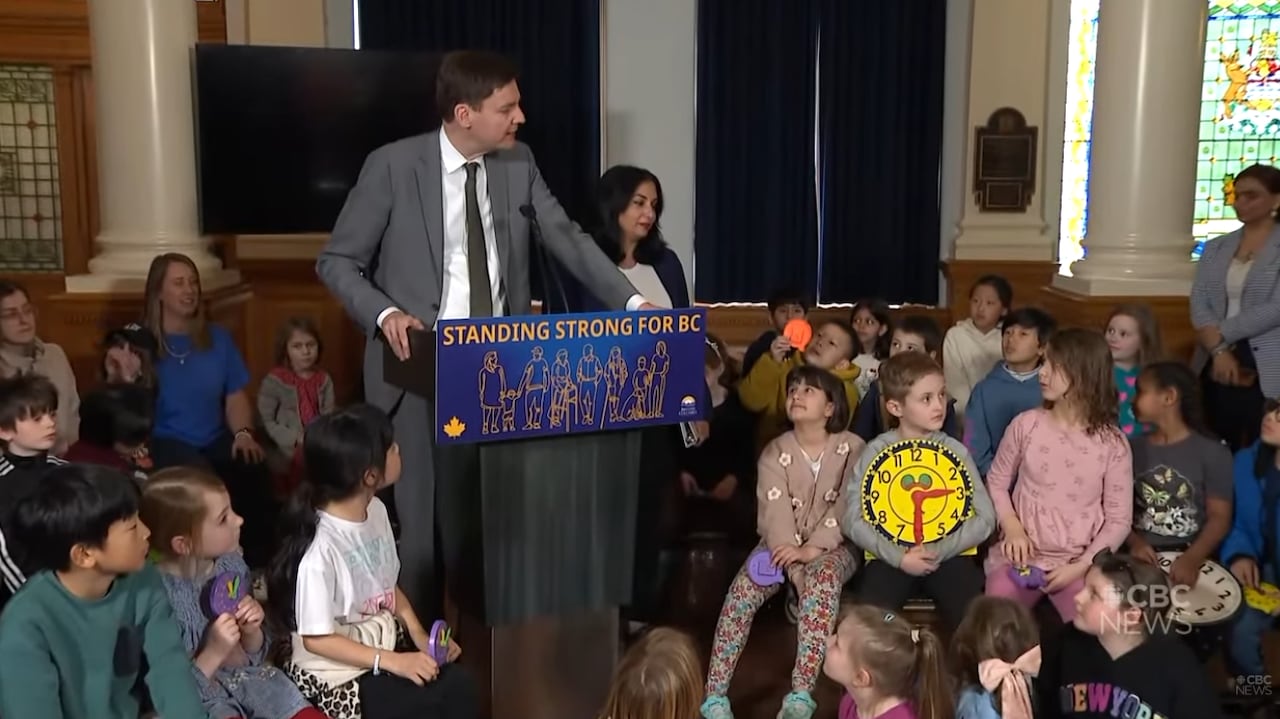 Premier David Eby at a podium in front of a group of school children. One boy holds a large kid's teaching clock.