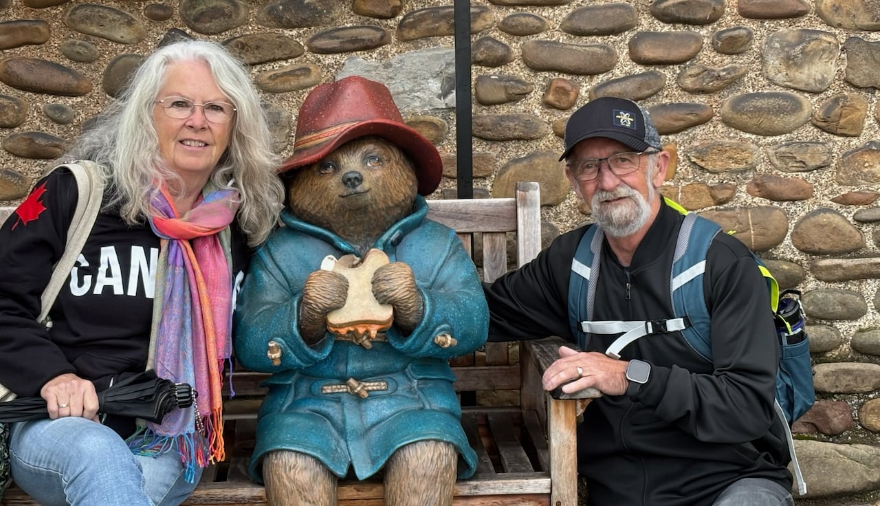 Man and woman, both with white hair, sit on a bench by a stone wall next to a statue of a bear wearing a blue jacket and a red hat