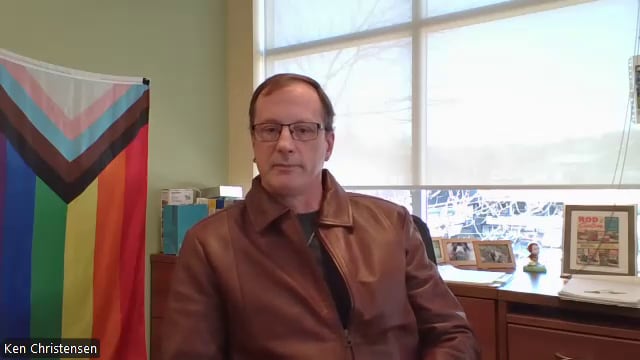 A man sits at a desk in a brown leather jacket and glasses 