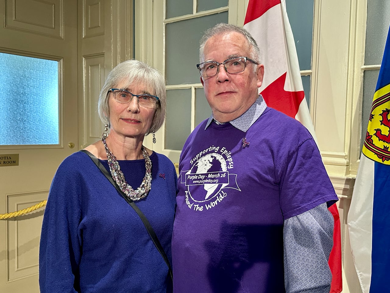 A woman with grey hair wearing a blue sweater stands to the left of a man with short grey hair who is wearing a purple shirt with the words, supporting epilepsy around the world, on it.