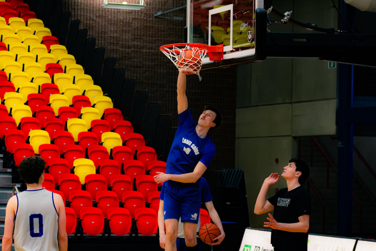 A tall man wearing blue dunks a basketball in the net as two others look on 