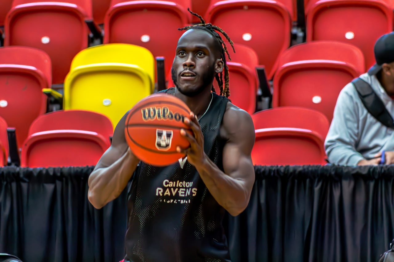 a Black man of about 20 in a dark short sleeved jersey holds a basketball in front of him appearing about to throw it