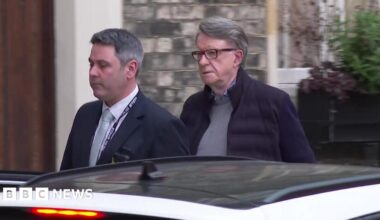 Lord Mandelson is led towards a car by a police officer who is not in uniform