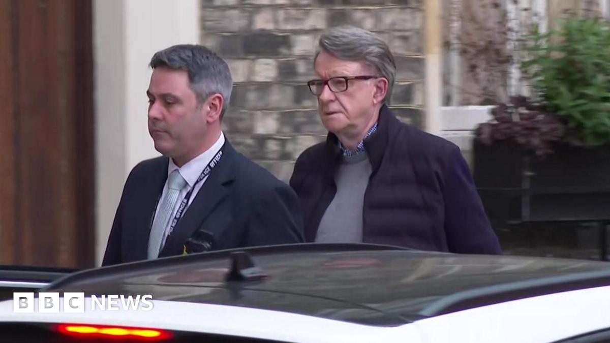 Lord Mandelson is led towards a car by a police officer who is not in uniform