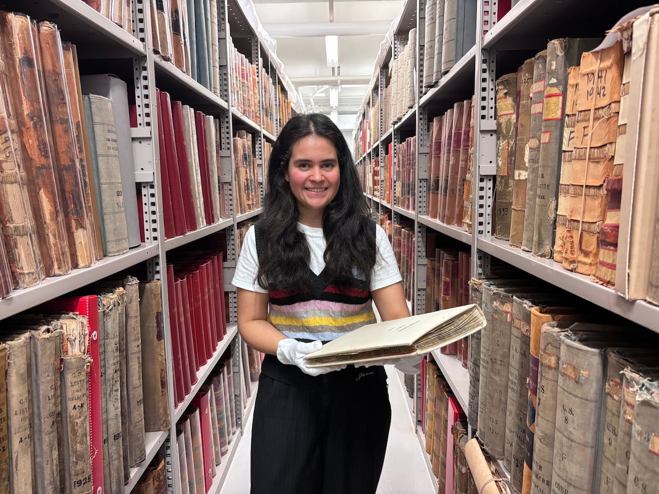 A woman wearing white gloves stands amid bookshelves filled with old registers.