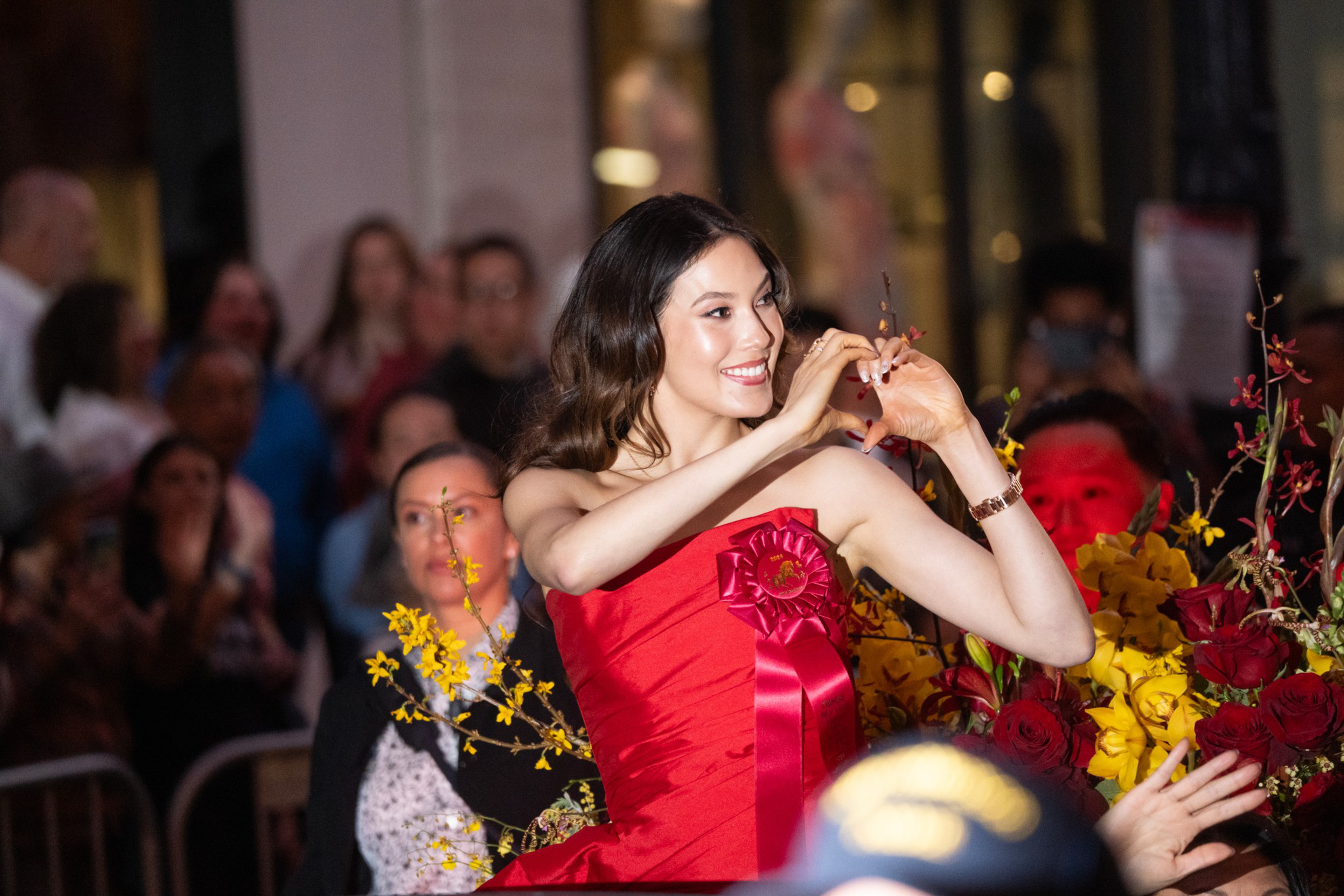 A woman in a red dress forms a heart with her hands while smiling, surrounded by red and yellow flowers and an applauding crowd in the background.