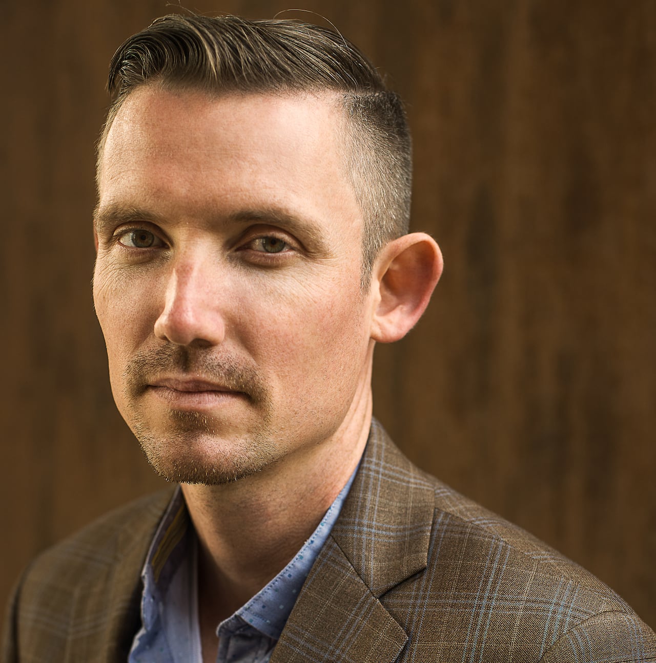 A man wearing a brown plaid blazer and blue shirt is shown in front of a brown background